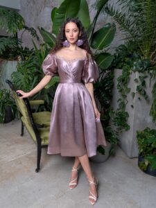 A woman is wearing a chic off-the-shoulder cocktail dress in a soft metallic hue, featuring voluminous puffed sleeves and a flattering pleated skirt, perfect for an elegant evening. Available by evening fashion store in Toronto.