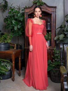 A woman is wearing a striking long-sleeve evening dress in a vibrant red hue, featuring elegant puffed sleeves and a pleated bodice with delicate embellishments at the neckline and waist. This refined design, ideal for a sophisticated evening look, is available by Papilio Boutique.