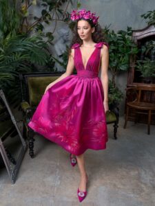 A woman is wearing a striking V-neck cocktail dress in a vibrant pink hue, featuring a pleated bodice and a full skirt with floral patterns. The elegant shoulder bow details add a playful touch, making it a perfect choice for special occasions. Available by evening dress store in Toronto.