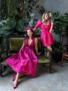 A woman is wearing a striking V-neck cocktail dress in a vibrant pink hue, featuring a pleated bodice and a full skirt with floral patterns. The elegant shoulder bow details add a playful touch, making it a perfect choice for special occasions. Available by evening dress store in Toronto.