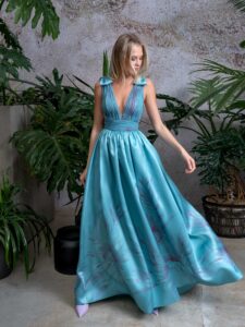 A woman is wearing a striking floor-length dress in a vibrant mint hue, featuring a pleated V-neck bodice and a full skirt with floral patterns. The elegant shoulder bow details add a playful touch, making it a perfect choice for special occasions. Available by evening dress store in Toronto.