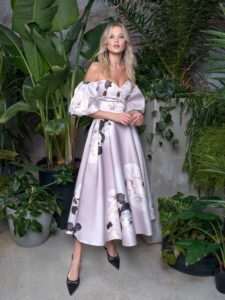 A woman is wearing an elegant off-the-shoulder cocktail dress with a voluminous skirt, in a soft lilac colour with floral print. The puff sleeves and fitted bodice add a touch of sophistication to this romantic ensemble, available by evening fashion store in Toronto.
