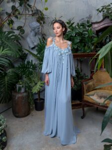 A woman is wearing a flowing chiffon maxi dress in a soft blue hue, featuring delicate floral appliqués along the neckline and cold-shoulder sleeves. This elegant and airy gown is perfect for formal events, available by evening dress store in Toronto.
