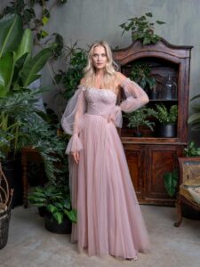 A woman is wearing a romantic tulle A-line evening dress in a soft blush tone, featuring delicate lace details on the bodice and voluminous off-the-shoulder sleeves. This elegant design is perfect for making a statement and is available by a Toronto Boutique of evening dresses.