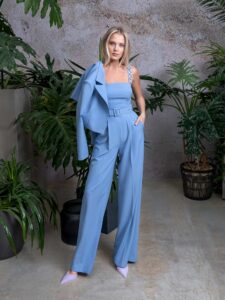 A woman is wearing a stylish sleeveless jumpsuit in light blue, paired with a matching cropped jacket and belt, showcasing a sleek and modern silhouette. Available by Papilio Boutique.