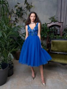 A woman is wearing a bright blue, mid-length evening dress with a full A-line skirt. It features a deep V-neck bodice adorned with delicate floral embroidery. An elegant look that’s available at an evening fashion store in Toronto.