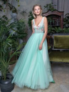 A woman is wearing a mint green floor-length evening dress. It features a full skirt and deep V-neck bodice adorned with delicate floral embroidery. This look is paired with a detachable long-sleeve tulle blouse with a neck bow. Available at an evening fashion store in Toronto.