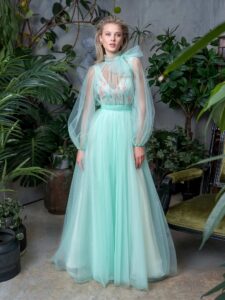 A woman is wearing a mint green floor-length evening dress. It features a full skirt and deep V-neck bodice adorned with delicate floral embroidery. This look is paired with a detachable long-sleeve tulle blouse with a neck bow. Available at an evening fashion store in Toronto.