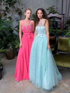 Two women are wearing floor-length tulle evening dresses, in vibrant pink and mint hues. They feature intricate floral detailing on the illusion-strapless bodices and flowing skirts, creating a graceful and elegant silhouette. Available at an evening fashion store in Toronto.