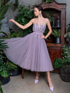 A woman is wearing a lavender tulle evening dress in midi-length. It features intricate floral detailing on the illusion strapless bodice and a flowing skirt, creating a graceful and elegant silhouette. Available at an evening fashion store in Toronto.