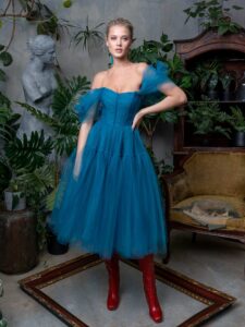 A woman is wearing a deep turquoise off-the-shoulder tulle ball gown with 3D flowers on the straps. It features a structured bodice and voluminous floor-length skirt. This elegant dress, available at a Toronto boutique of evening dresses, is perfect for making a bold statement.