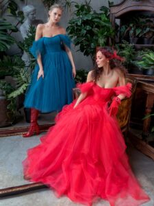 A woman is wearing a bold red off-the-shoulder tulle ball gown with 3D flowers on the straps. It features a structured bodice and voluminous skirt in midi-length. t. This elegant dress, available at a Toronto boutique of evening dresses, is perfect for making a bold statement.