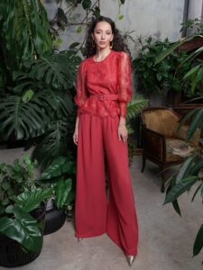 A woman is wearing a two-piece set available from an evening dress store in Toronto. This striking ensemble features a cropped lace top with a Peter Pan collar and puffed long sleeves, paired with high-waisted, flowy pants in a matching red hue, offering a chic and modern twist on traditional evening wear.