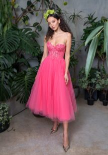 A woman standing in a verdant indoor garden, wearing a hot pink prom dress with intricate bodice embroidery and a yellow floral headpiece, paired with metallic pointed-toe heels, representing stylish prom dresses Toronto.