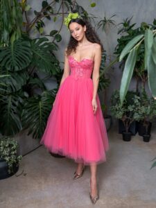 A woman is wearing a spaghetti strap ballgown available from an evening fashion store in Toronto. This vibrant pink gown features a delicate, floral-embroidered bodice with sheer details, flowing into a voluminous tulle skirt in midi-length that adds a whimsical and romantic touch to the ensemble.