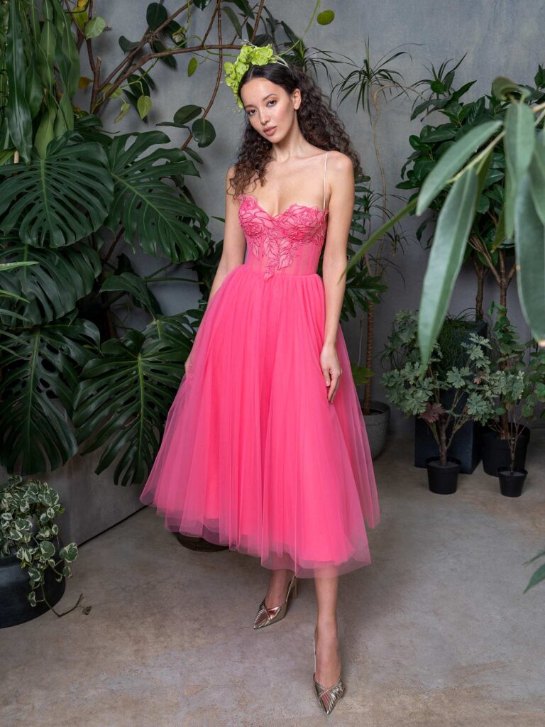 A woman standing in a verdant indoor garden, wearing a hot pink prom dress with intricate bodice embroidery and a yellow floral headpiece, paired with metallic pointed-toe heels, representing stylish prom dresses Toronto.