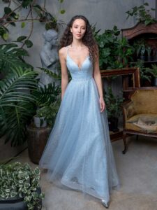 A woman is wearing a light blue A-line evening dress with spaghetti straps available from Papilio Boutique. This elegant dress features a plunging neckline with a sheer inset and a shimmering fabric overlay, creating a sophisticated and timeless look.