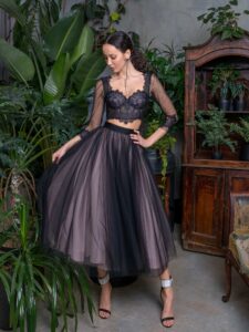 A woman is wearing a delicate two-piece set in black available from Papilio Boutique, featuring a lace crop top with sheer three-quarter sleeves and a voluminous tulle skirt. This elegant ensemble offers a modern twist on classic evening wear.