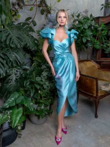 A woman is wearing a stunning strapless cocktail dress in a vibrant aqua color, accented with dramatic ruffled shoulders and a belted waist, available from an evening fashion store in Toronto. This dress is perfect for making a bold statement at any upscale event.