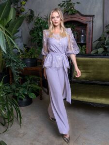 A woman is wearing an elegant blouse and wide-leg pants set in a soft lavender hue, featuring delicate lace details and a belted waist, available from an evening fashion store in Toronto. Perfect for a sophisticated and modern evening look.