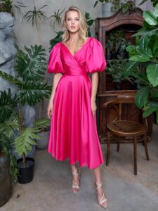 A woman is wearing a vibrant pink Satin A-line cocktail dress with short puff sleeves and a pleated V-neckline. This elegant ensemble dress is paired with white strappy sandals and is available from a Toronto boutique of evening dresses.