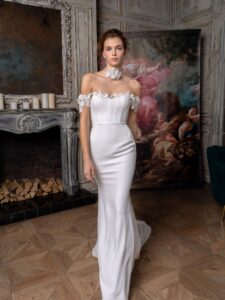 A striking fit-and-flare wedding dress from Papilio's Toronto bridal boutique is modeled, showcasing off-the-shoulder lace sleeves and a slimming silhouette, set against an ornate fireplace and antique mirror.
