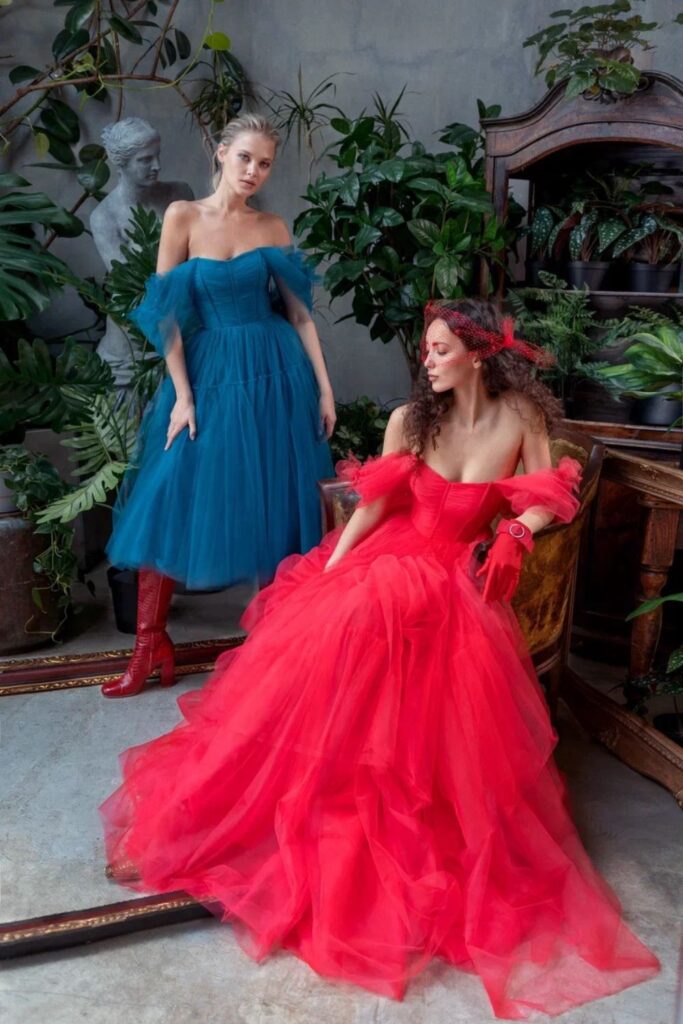 Two women posing in a botanical backdrop, one standing in an off-the-shoulder rich blue tulle dress and red boots, the other seated in a voluminous bright red tulle dress with off-the-shoulder ruffles, showcasing ballgown prom dresses Toronto.