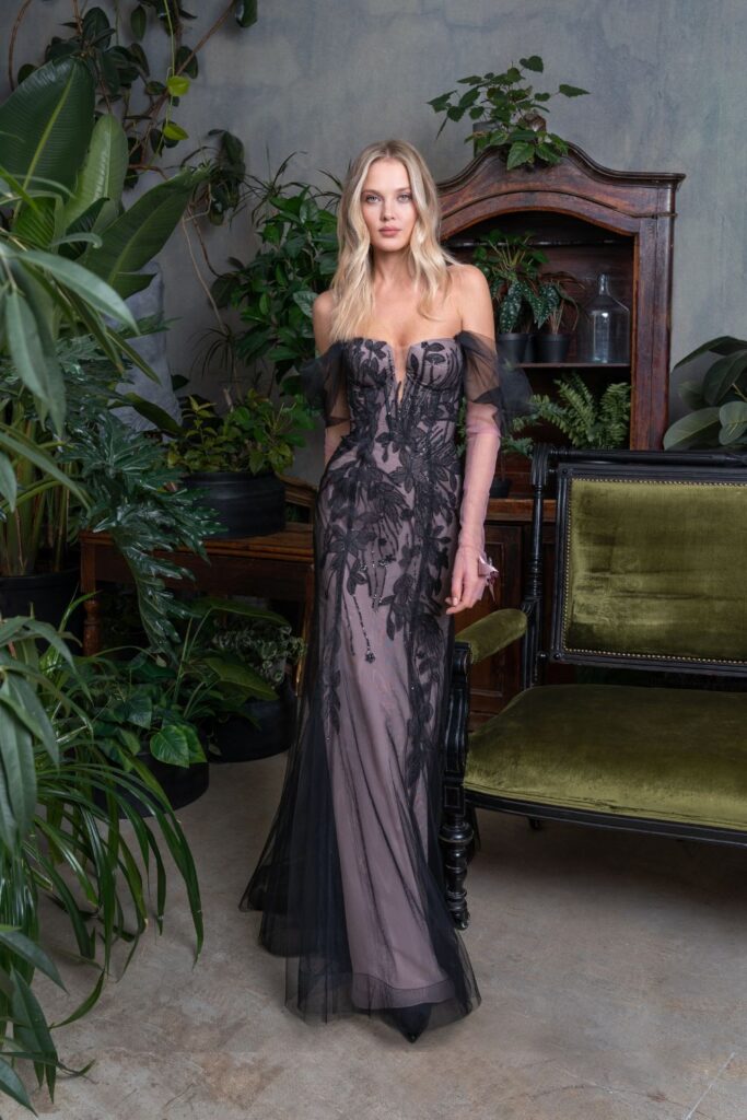 A woman standing amidst indoor plants, wearing an elegant fitted prom dress in black with transparent overlay and floral embroidery, exemplifying the sophistication of fitted prom dresses Toronto.