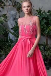 A woman in a botanical setting wearing a coral pink prom dress with a sheer overlay bodice, adorned with delicate floral appliqués and a satin bow at the waist, representing pink prom dresses Toronto.