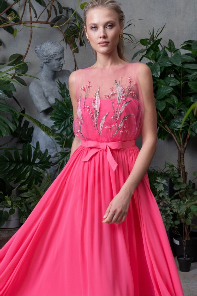 A woman in a botanical setting wearing a coral pink prom dress with a sheer overlay bodice, adorned with delicate floral appliqués and a satin bow at the waist, representing pink prom dresses Toronto.