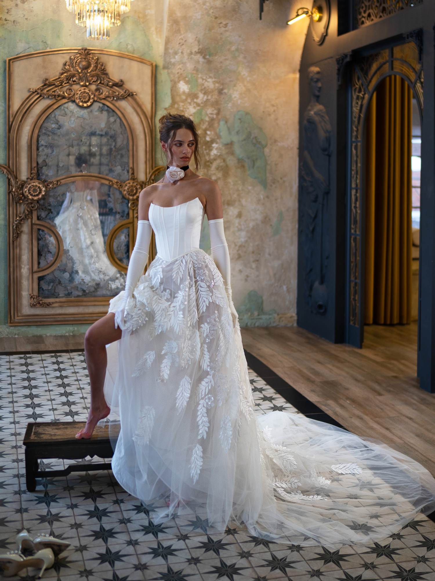 A model poses in an opulent room wearing a contemporary strapless wedding dress with long sleeves and detailed white leaf embroidery on the skirt and train, highlighting a unique wedding dress style by Papilio Boutique.