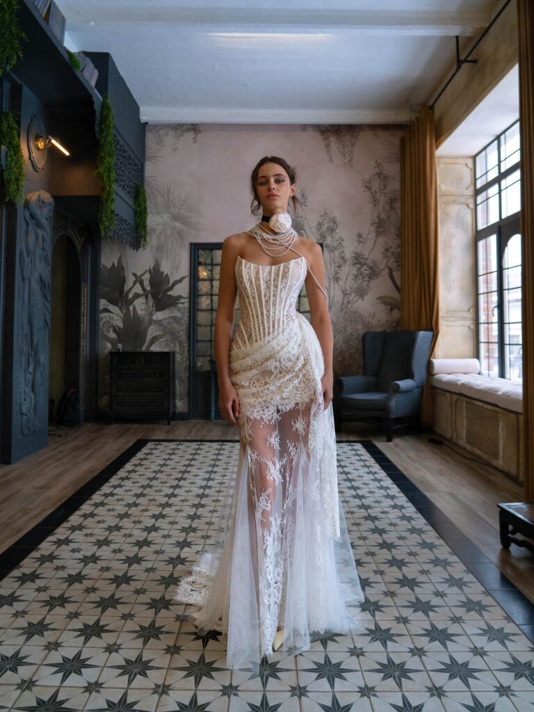 A model stands gracefully in a sophisticated setting wearing a sleeveless wedding dress with intricate white lace and beadwork, a corset bodice, and a sheer lace skirt, exemplifying an elegant wedding dress style by Papilio Boutique.