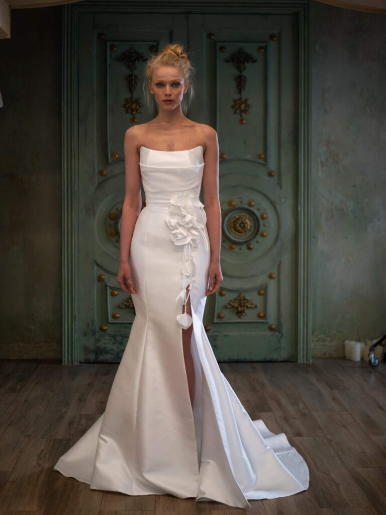 A woman trying on a wedding dress by Papilio Boutique with a strapless design and a dramatic thigh-high slit, adorned with a cascading side flower, stands against an antique turquoise door, contemplating her reflection during her bride dresses fitting.