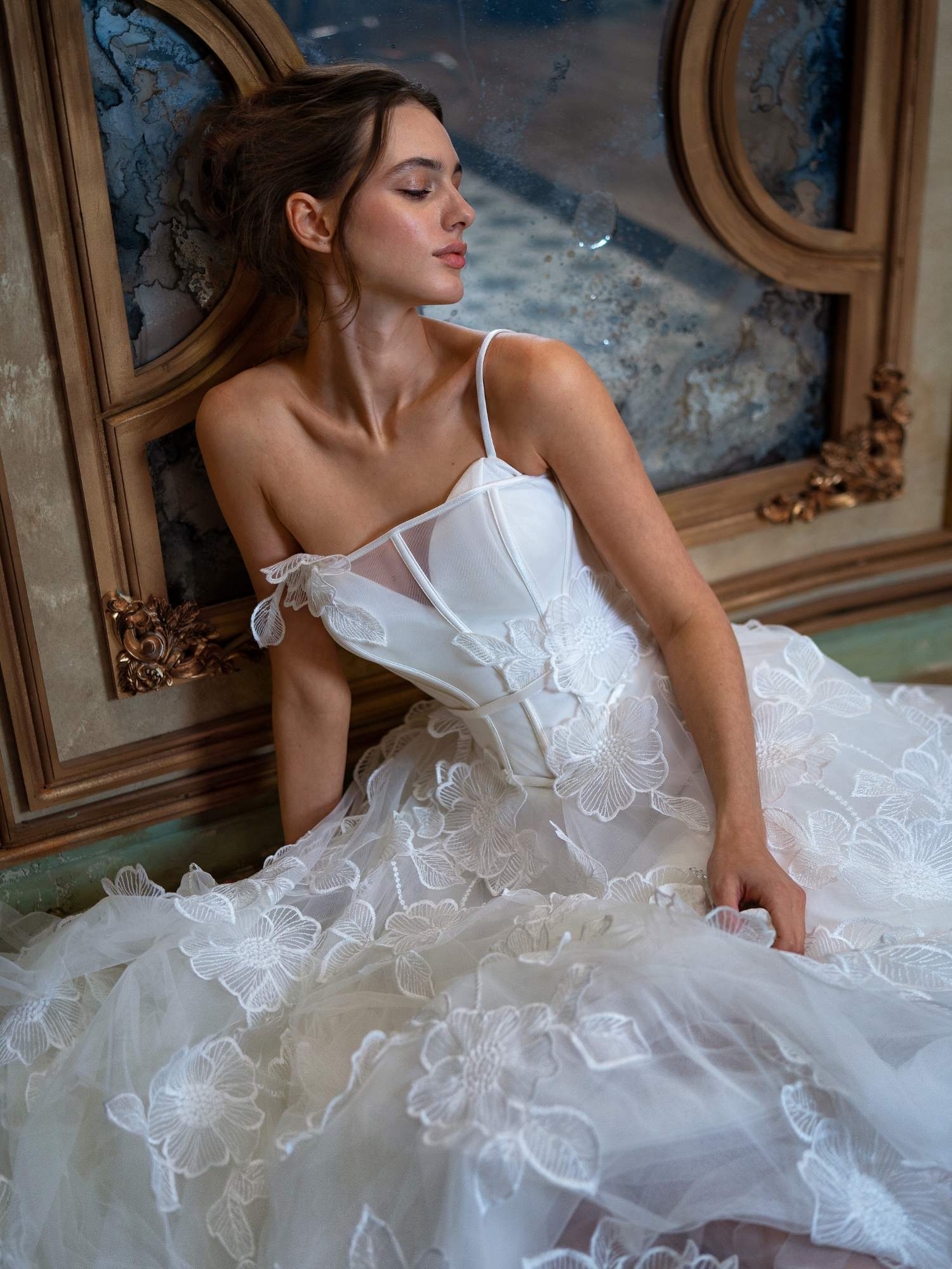A serene bride gazes downward while seated, draped in an ethereal white gown with delicate floral embroidery, similar to one of the exquisite finds at Papilio's sample sale in Toronto for wedding dresses, surrounded by a room rich in vintage charm and ornate details.