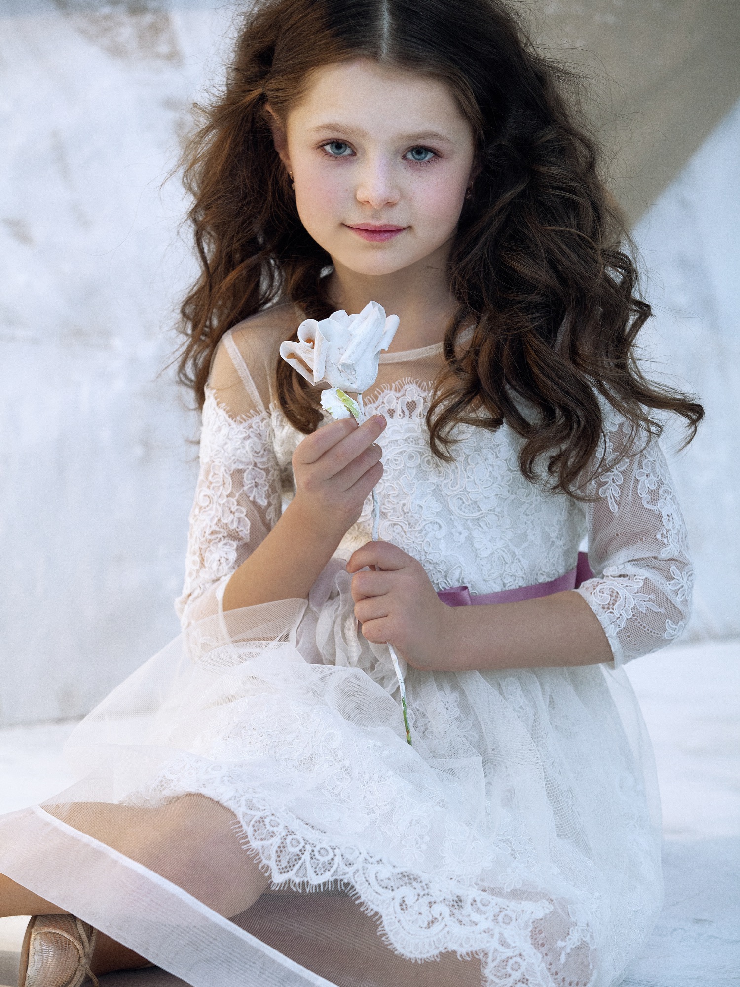 Young girl wearing a lace baptism dress Toronto, featuring sheer sleeves and floral details, holding a white flower while seated in a soft, elegant setting.