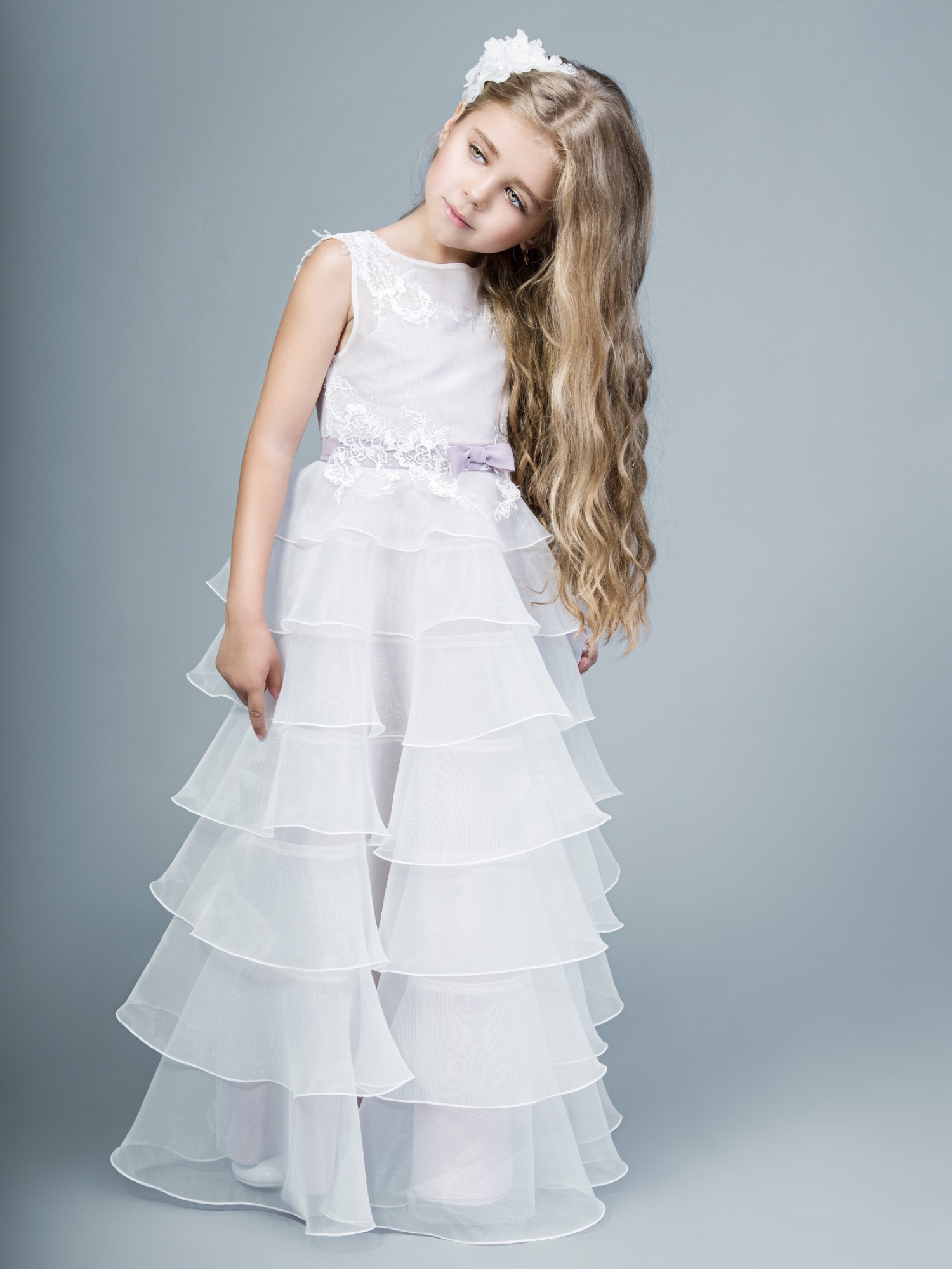 Young girl wearing a tiered white gown with lace bodice and lavender bow, styled with a floral hair accessory – first communion dresses Toronto.