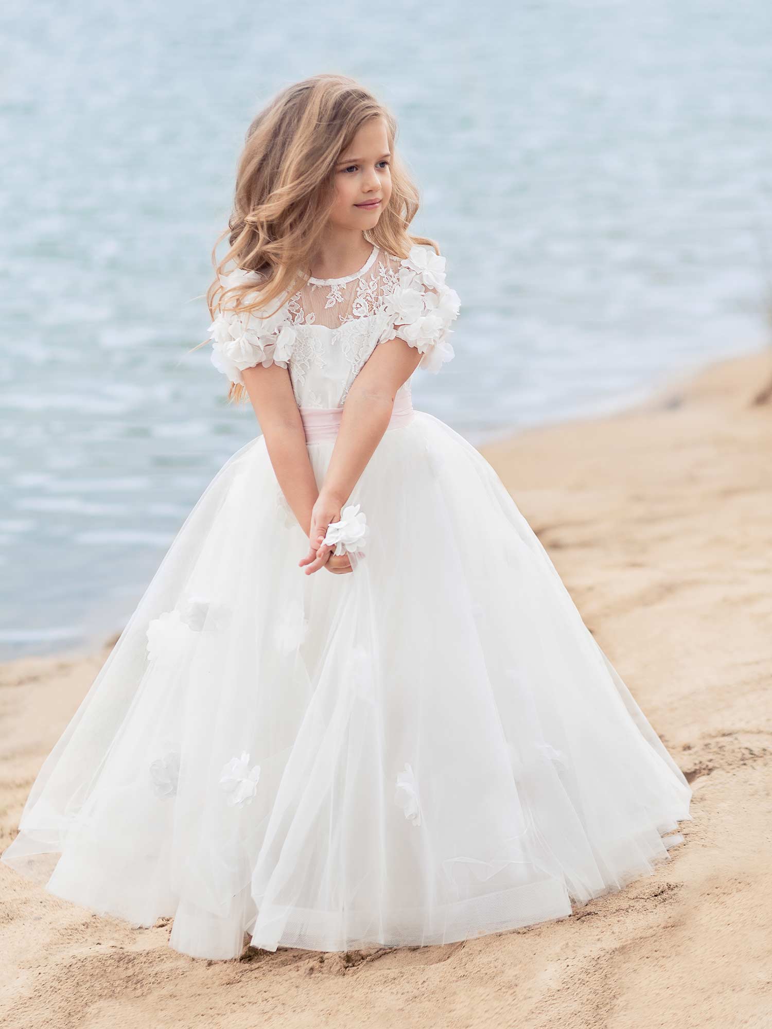 Young girl in an elegant white tulle gown with floral appliqué sleeves and pink sash, standing on the beach – girls formal dresses Toronto.