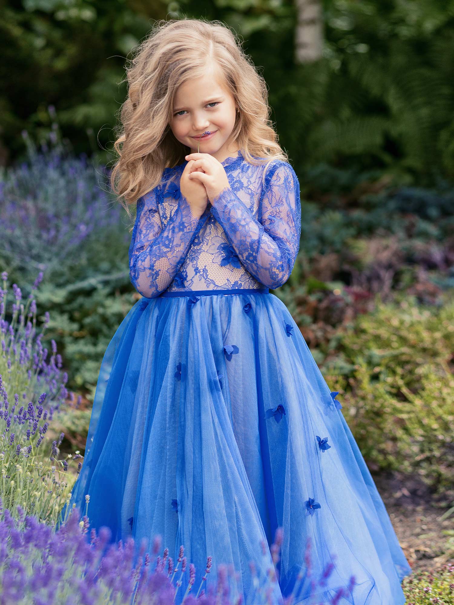 Smiling young girl wearing a royal blue lace and tulle gown with floral appliqués, posing outdoors among lavender fields – girls formal dresses Toronto.